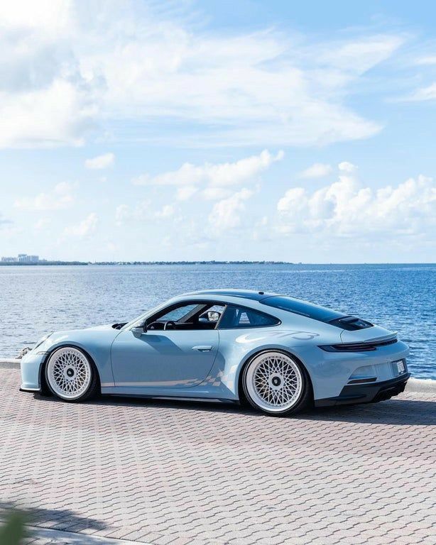 Sleek silver sports car parked by the ocean under a blue sky, showcasing striking design and custom wheels.