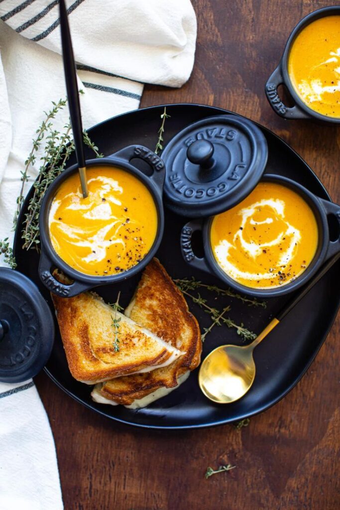 Creamy pumpkin soup in bowls with grilled cheese sandwiches on a black plate.