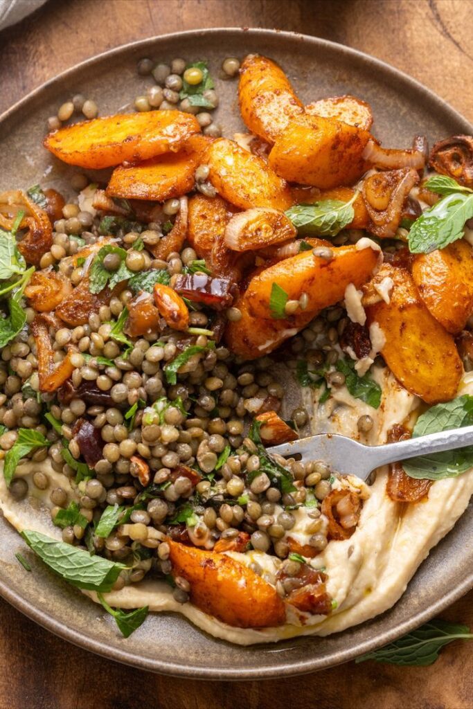 Roasted carrots and lentils with herbs on creamy hummus, garnished with fresh mint leaves on a round plate.