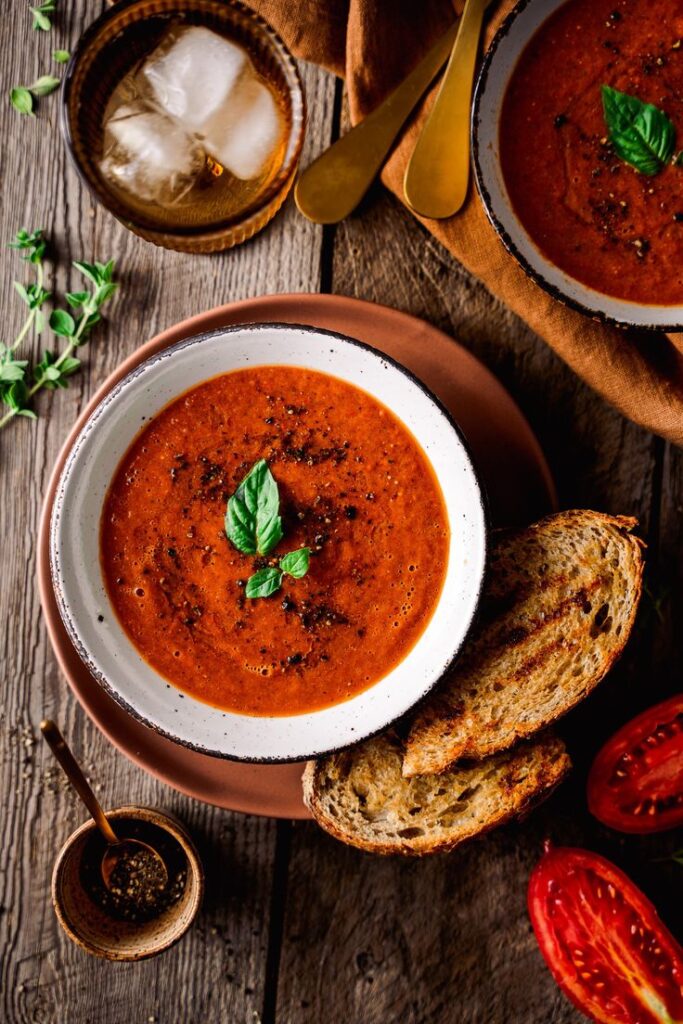 Rustic tomato soup with basil garnish served with crusty bread on a wooden table.