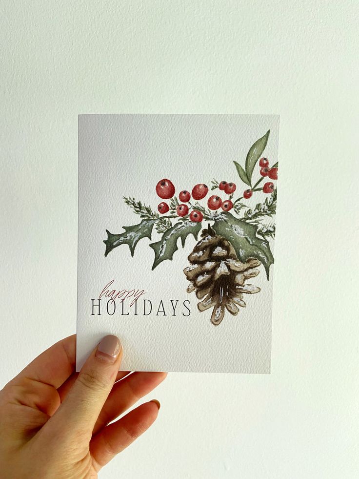 Hand holding a festive card with Happy Holidays text, featuring pinecones and holly berries on a white background.