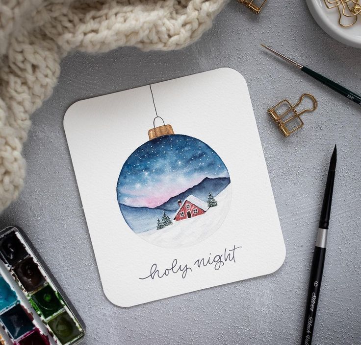 Watercolor painting of a winter scene with a cabin in an ornament, captioned holy night, surrounded by art supplies.