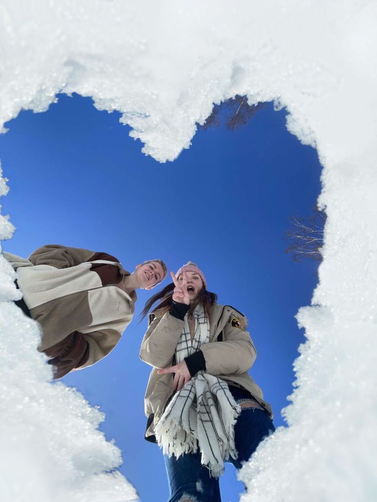 Two people enjoying a winter day, seen through a heart-shaped snow frame against a bright blue sky.