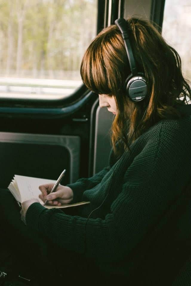 Woman with headphones writing in a notebook on a bus journey.