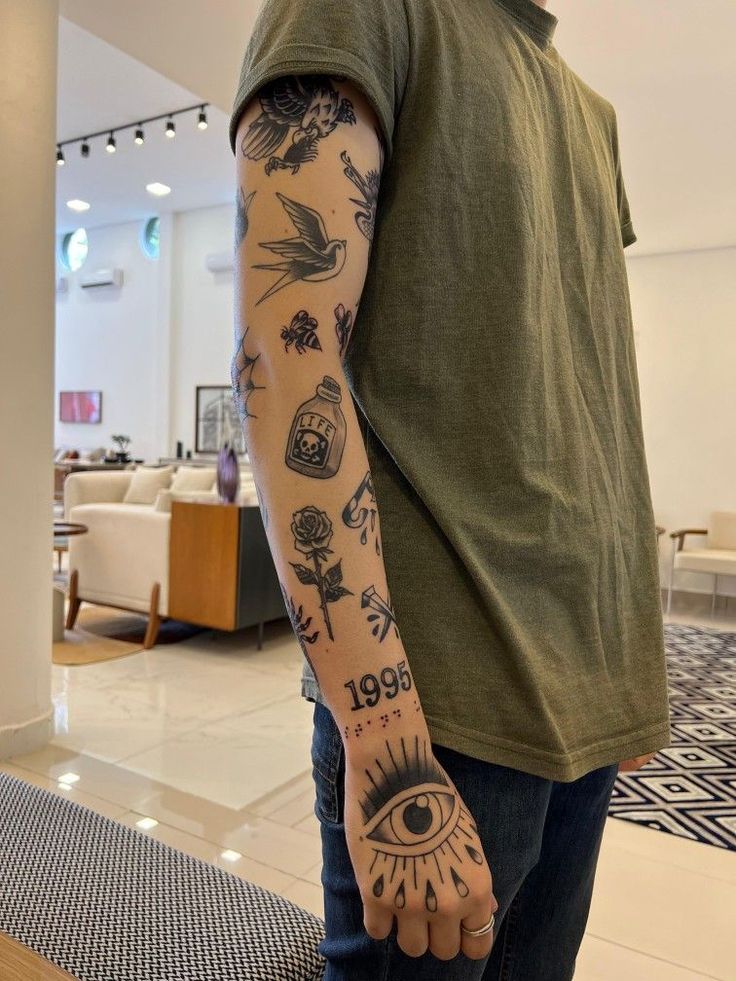 Arm with various black tattoos including a dove, rose, 1995, and an eye design, wearing olive t-shirt.