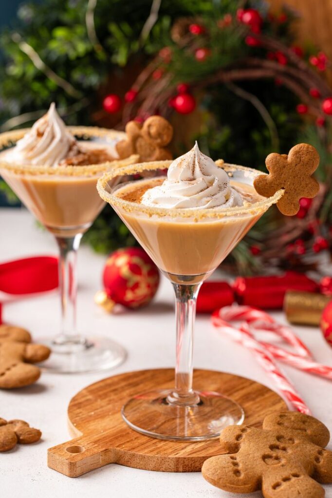 Festive gingerbread martinis with whipped topping and cookies on glass rims, surrounded by Christmas decor.