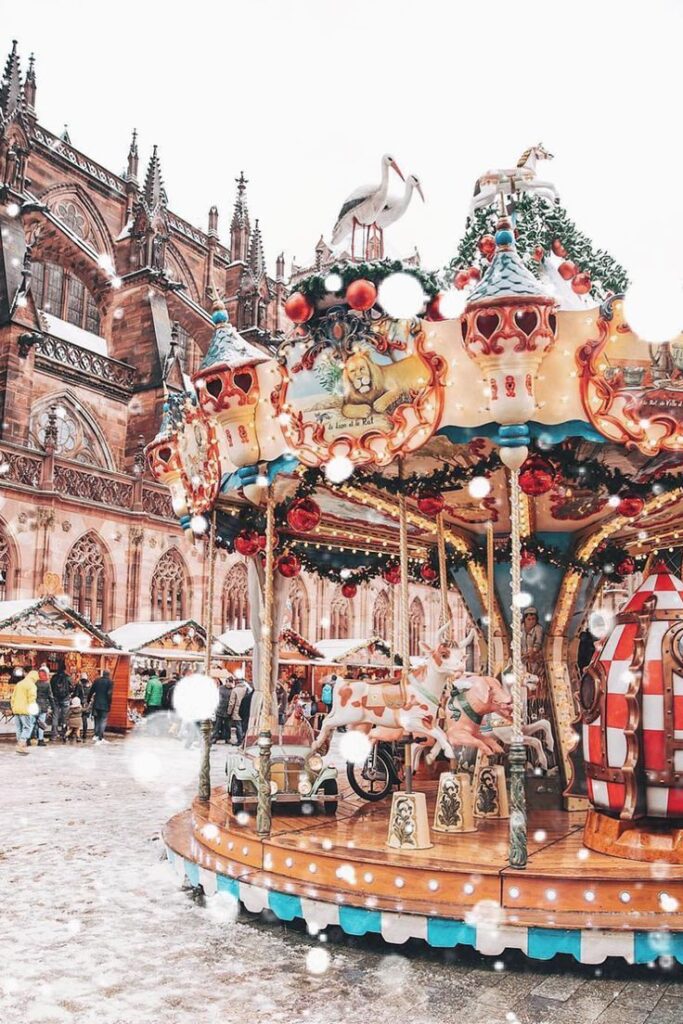 Festive carousel in front of cathedral with snowflakes, surrounded by Christmas market stalls and decorations.