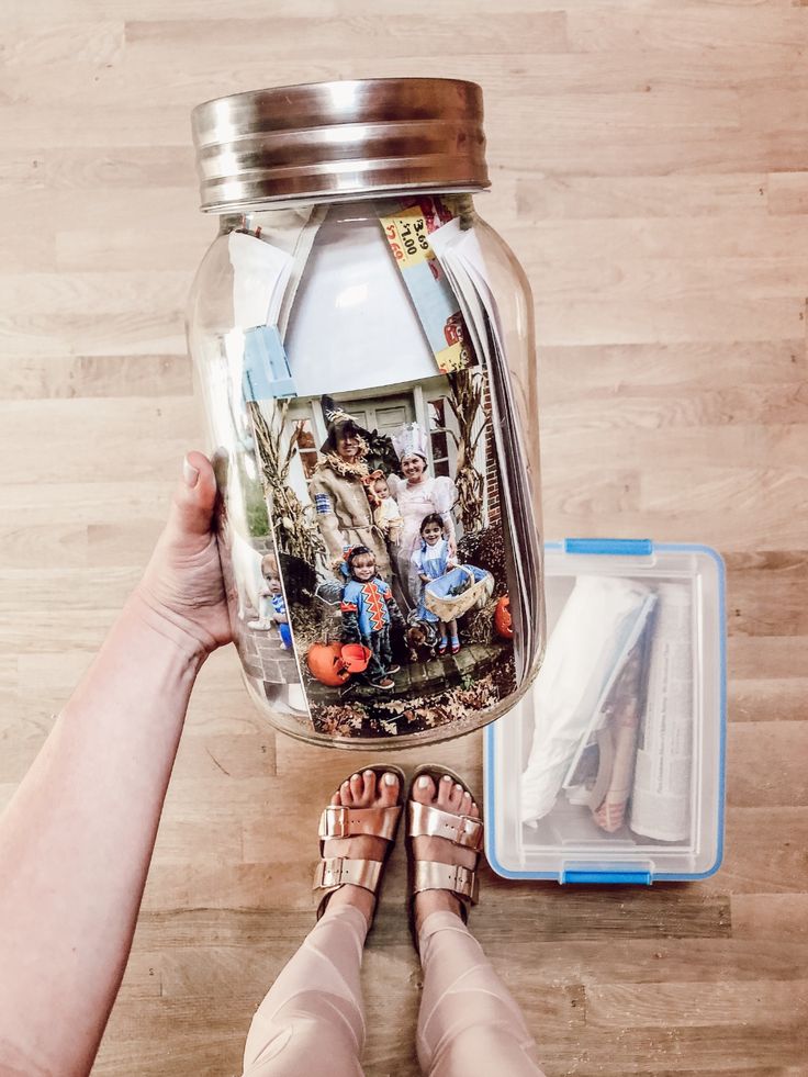 Person holding a memory jar filled with photos and keepsakes, standing on a wooden floor near a blue storage box.