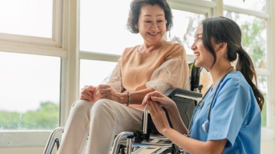 Caregiver smiling with elderly woman in wheelchair near a sunny window, providing supportive and compassionate care.