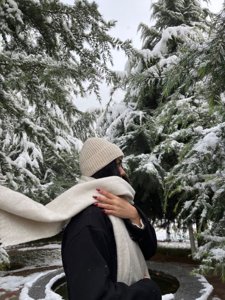 Woman in winter clothing amidst snowy pine trees, wearing beige hat and scarf. Serene winter scene.