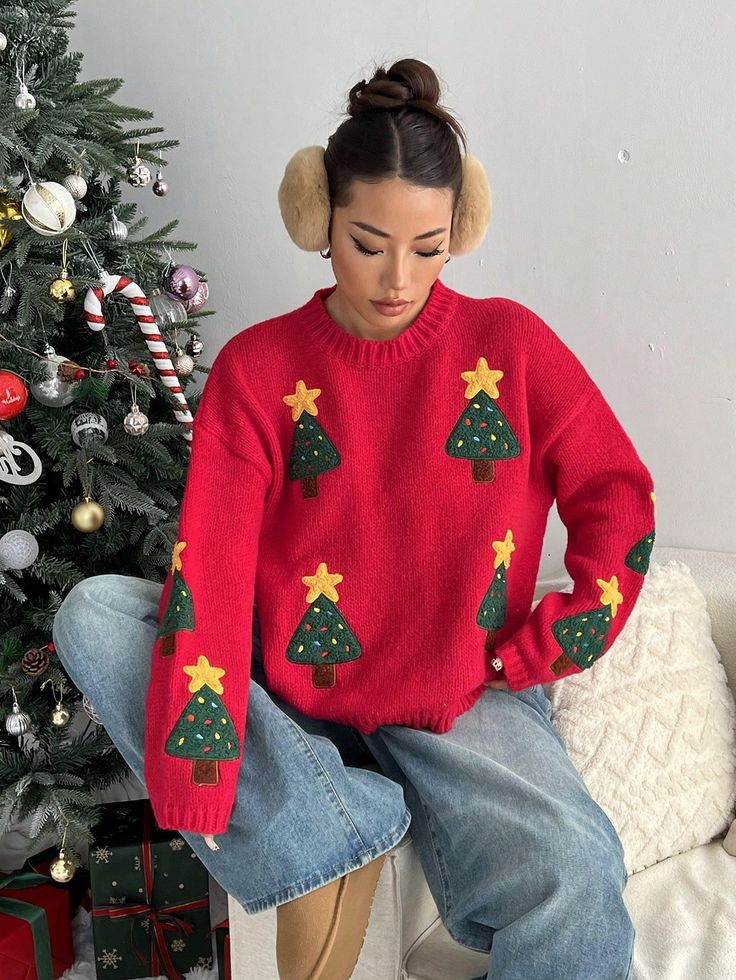 Woman in festive red Christmas sweater with tree design, sitting by a decorated tree, wearing earmuffs and jeans.