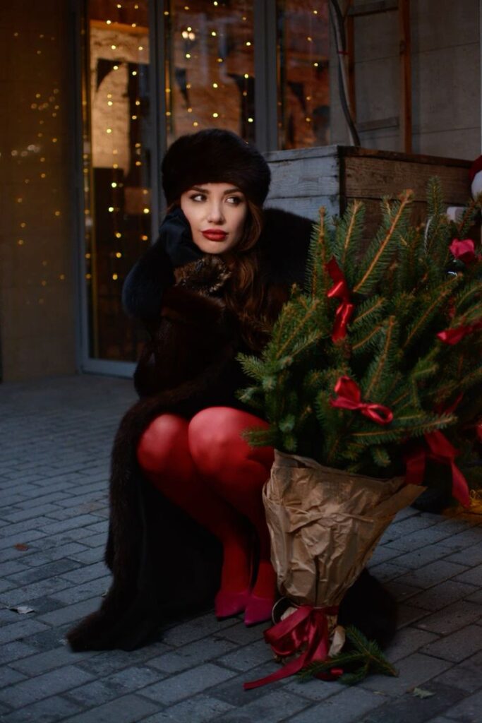 Woman in winter outfit with red tights sits beside wrapped small Christmas tree on a festive, illuminated street.