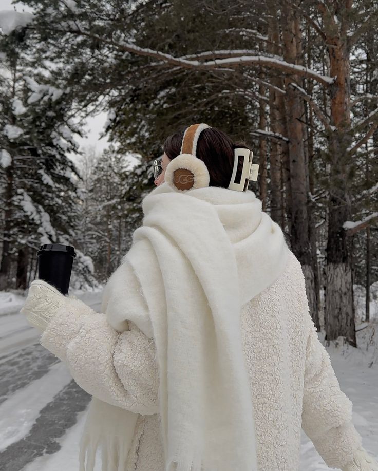 Person in winter attire holding a coffee cup, walking in a snowy forest, wearing earmuffs and a fluffy coat.