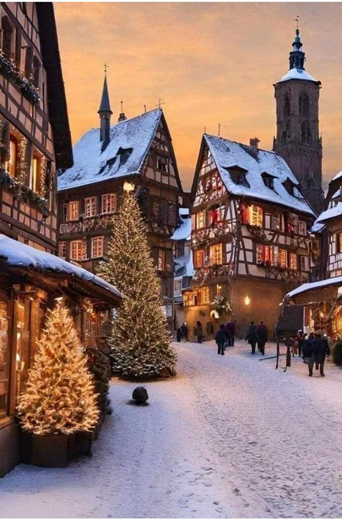 Charming snowy village street with lit Christmas trees, traditional houses, and church tower at sunset.