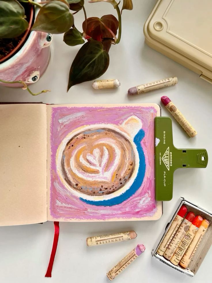 Open sketchbook with latte art drawing surrounded by pastel crayons and plant.