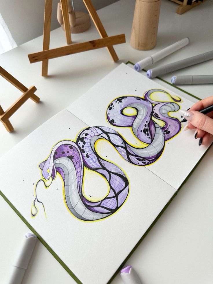 Artistic snake sketch in a notebook, featuring purple and yellow hues, with markers on a white desk.