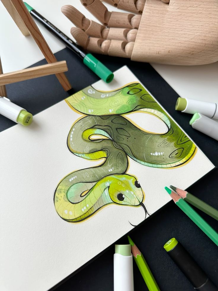 Green snake illustration with markers, pencils, and a wooden hand model on a black background.