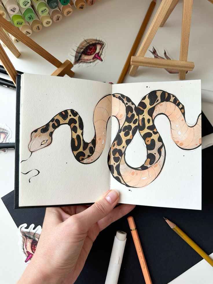 Hand holding sketchbook with colorful snake drawing, surrounded by art supplies on a table.