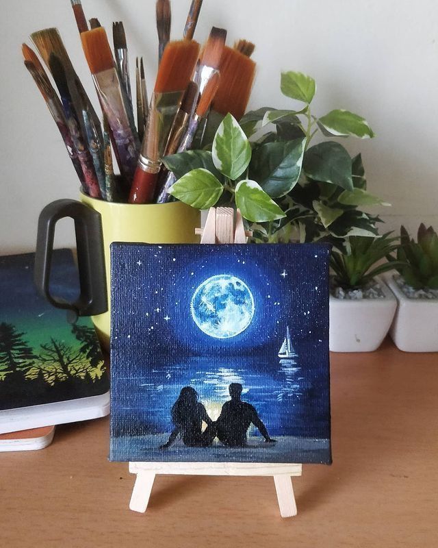 Miniature canvas painting of a couple by moonlit sea, with brushes and plants in the background.