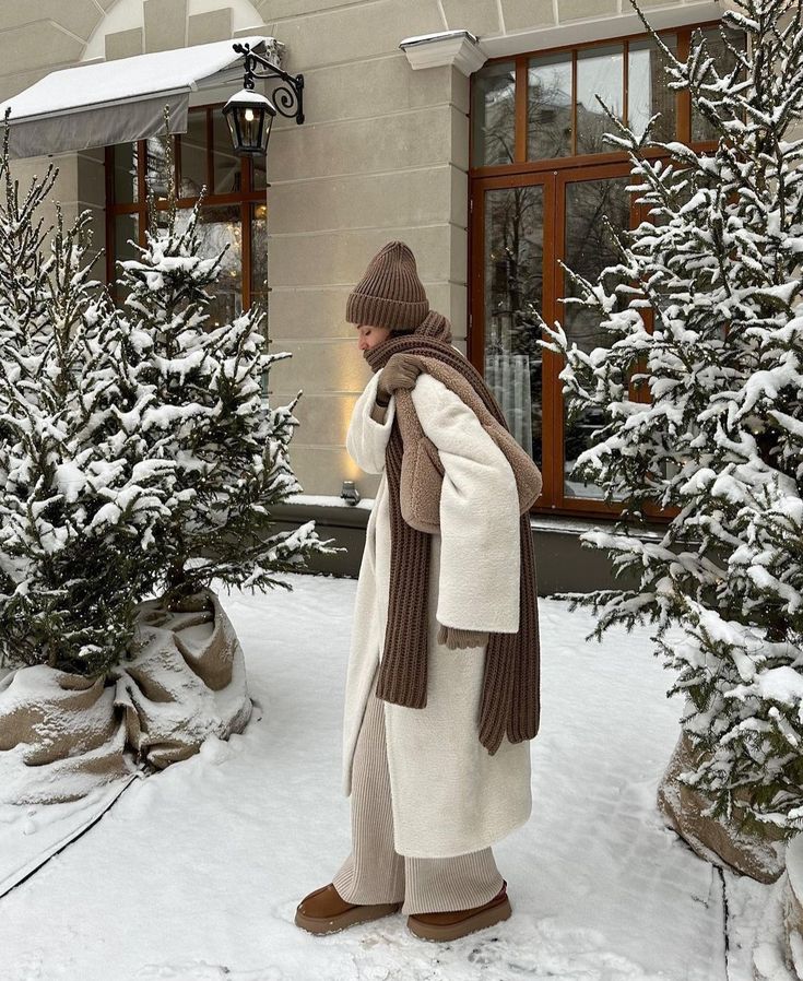 Person in cozy winter attire walking through snowy garden near elegant building.