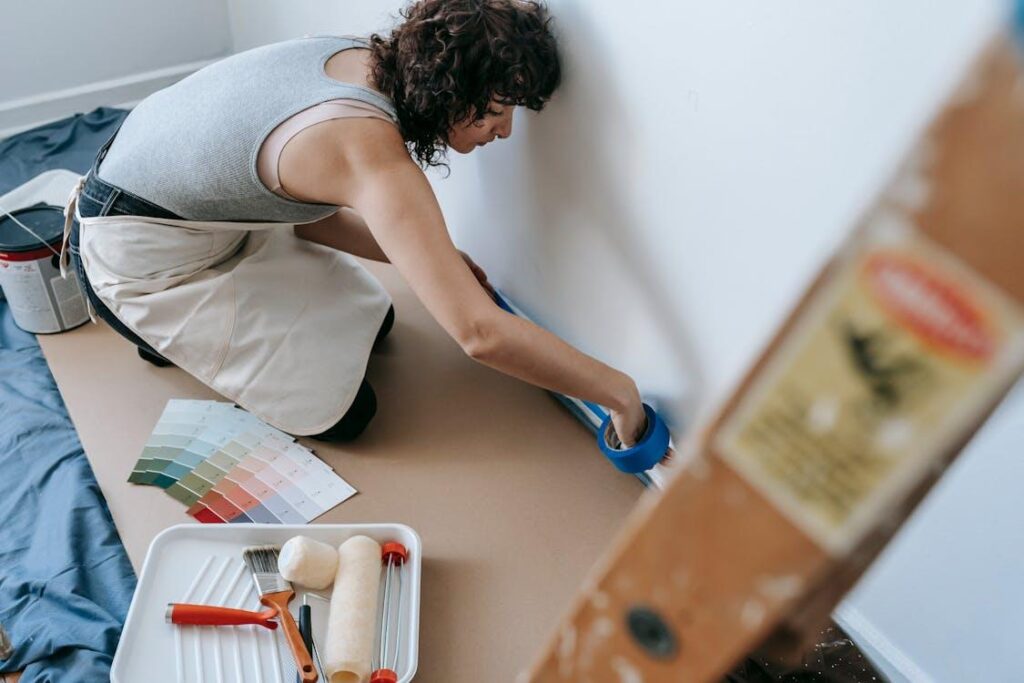 Person preparing to paint a wall with tape, paint palette, roller, and brush nearby on a floor.