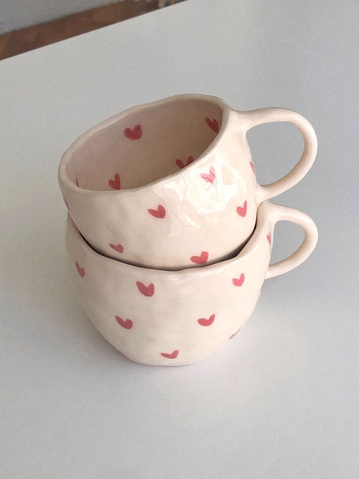 Two stacked ceramic mugs with pink heart patterns on a white table.