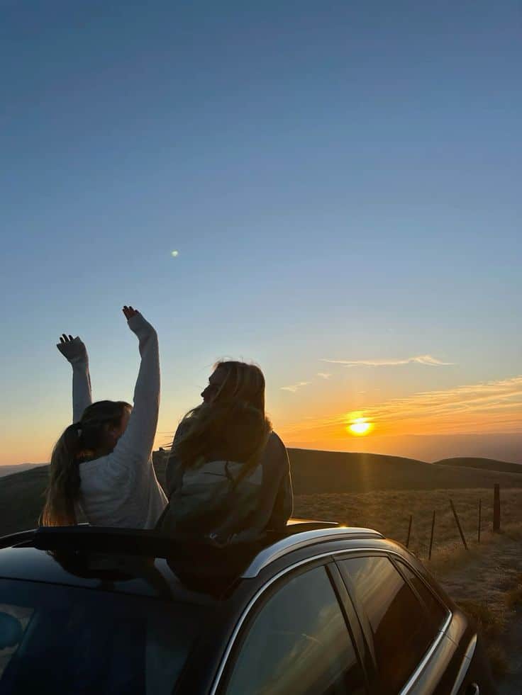 Two friends enjoy sunset from a car roof, with scenic hills and glowing horizon in the background.