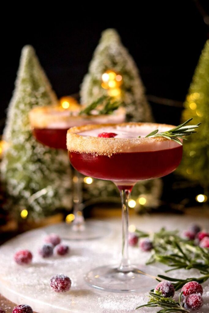 Festive holiday cocktail with rosemary garnish, sugar rim, and cranberries, set against a background of lit Christmas trees.