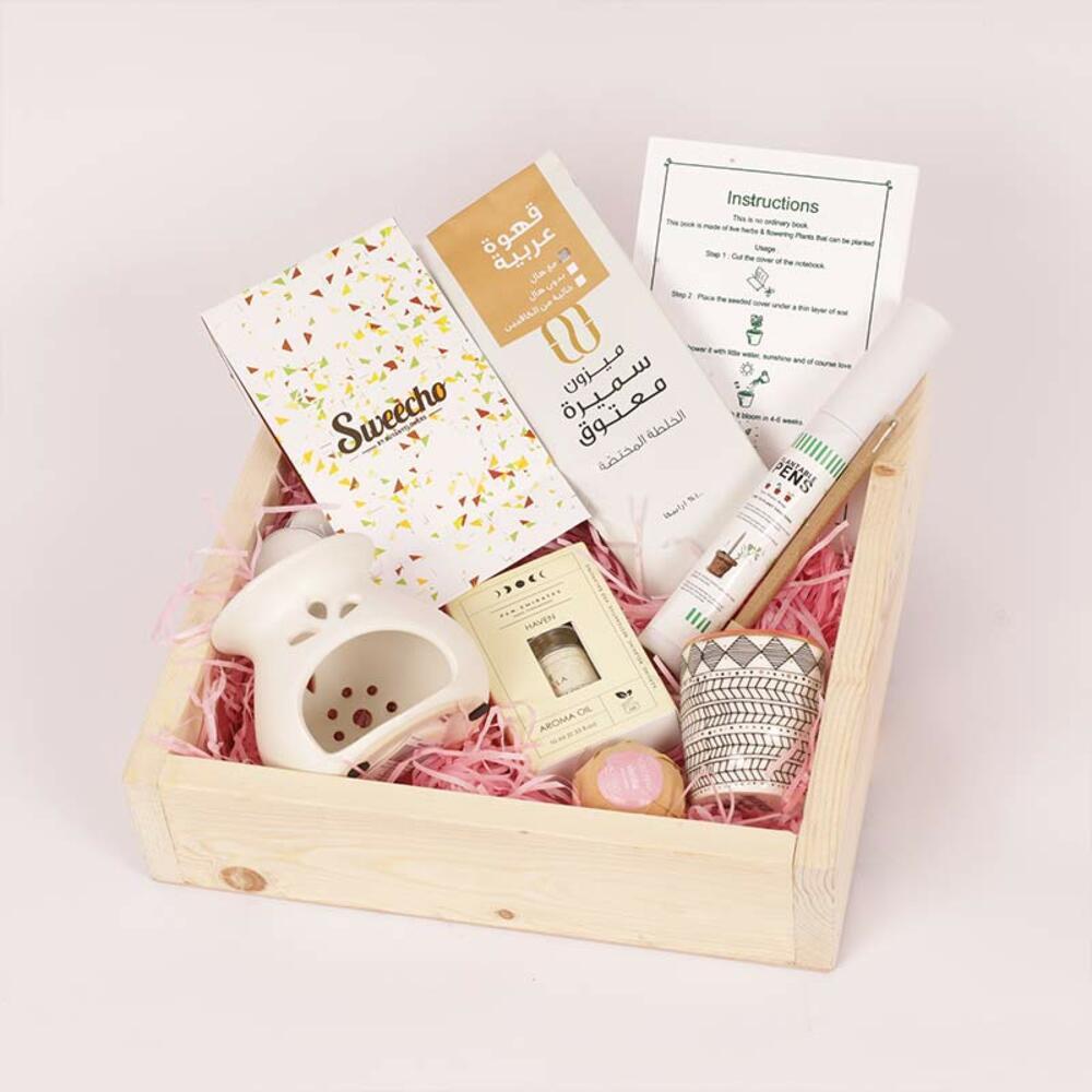 Gift box with Arabic coffee, aroma oil, art cup, and thoughtful items on pink filler in a wooden crate.