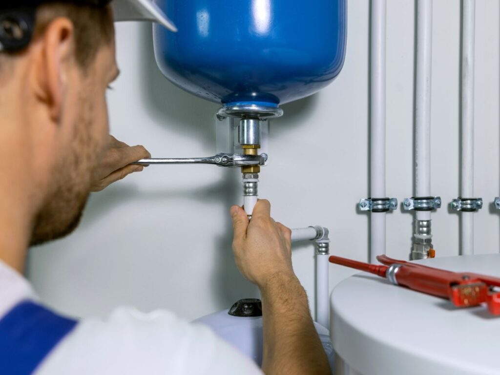 Plumber using wrench to install water heater in utility room, focusing on blue tank and connected pipes.