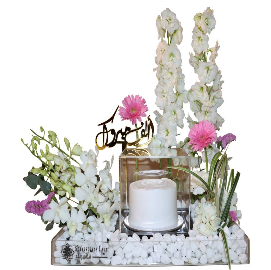 Elegant floral arrangement with white and pink flowers, white pebbles, and a small white cake in a glass display.