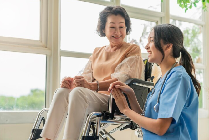 Caregiver in blue uniform smiling at woman in wheelchair by large sunny window.