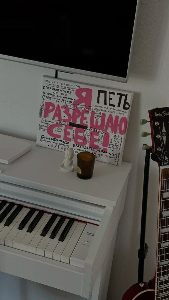 White piano and guitar in a room with Russian text art and a small candle on top.