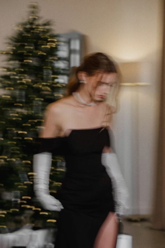 Elegant woman in black dress and gloves walking past Christmas tree in a softly blurred setting.