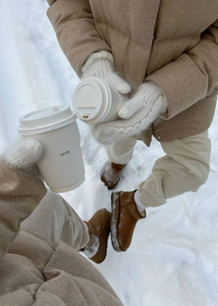 Two people in beige winter coats, holding coffee cups in the snow. Cozy winter scene.