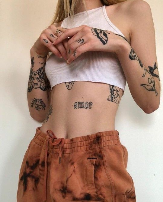 Woman with body tattoos, wearing a white crop top and orange pants, showcases unique tattoo art and style.