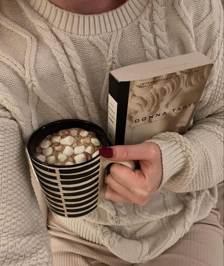 Cozy scene with a person in a sweater holding hot chocolate and a book, creating a perfect reading moment.