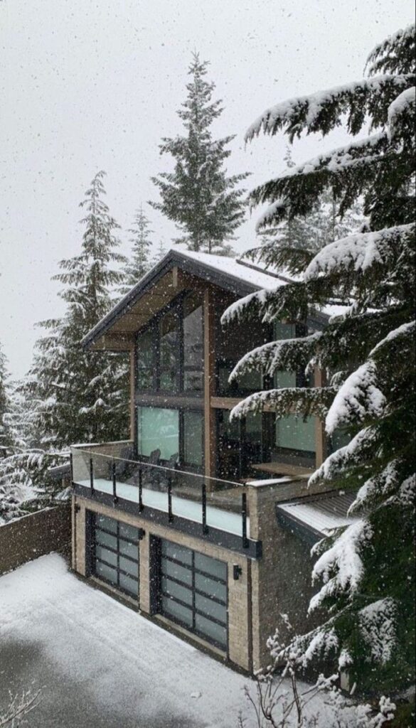 Modern mountain cabin surrounded by snow-covered trees during winter snowfall.