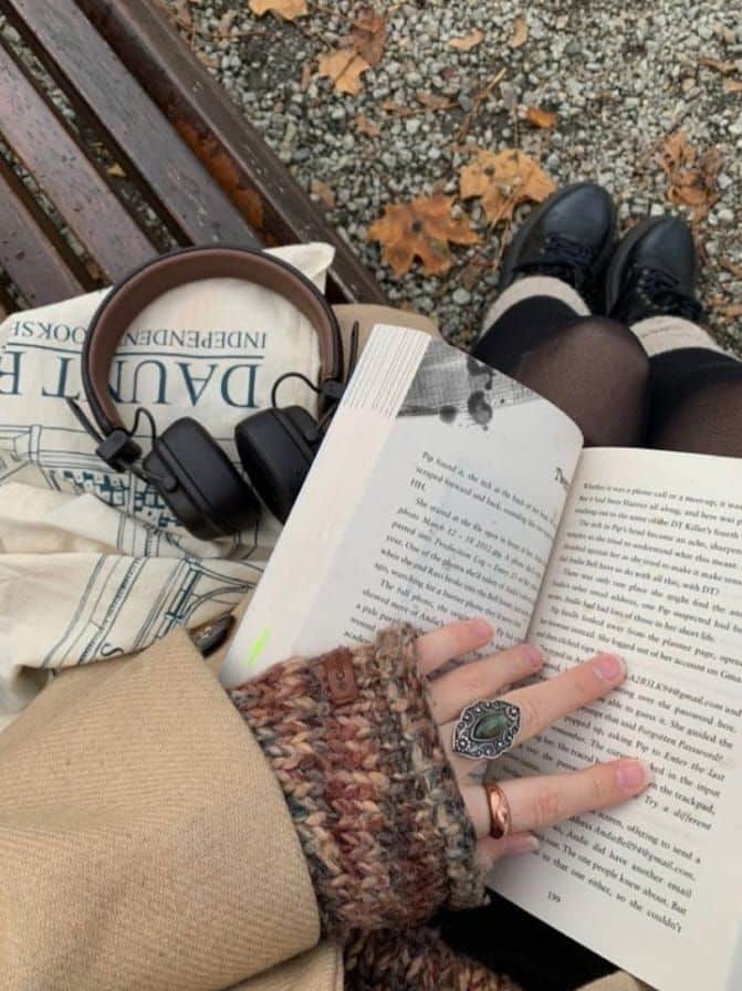Cozy autumn reading with open book, knitwear, headphones, and fallen leaves on a park bench.