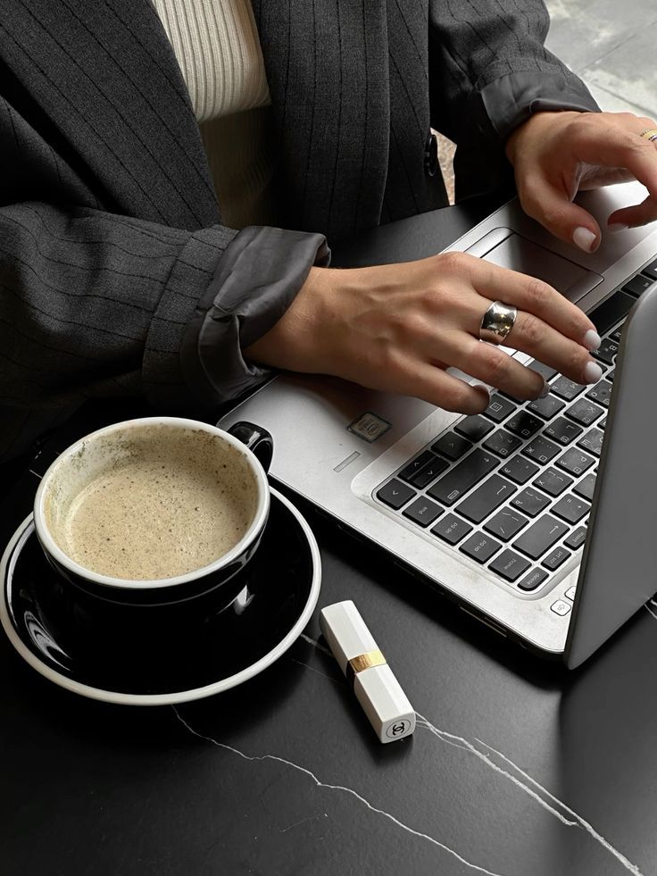 Businesswoman typing on laptop with coffee and lipstick on desk.