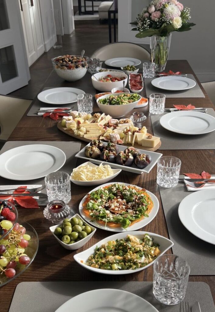 Elegant dining table set for a feast with salads, cheese platter, fruits, and floral centerpiece.