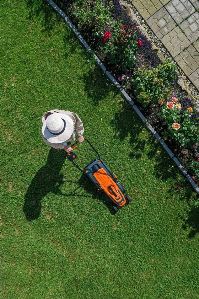 Person mowing grass with an orange lawn mower in a garden with colorful flowers, view from above.