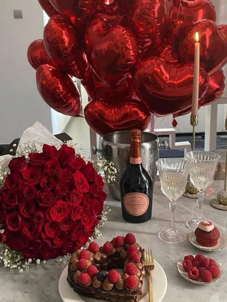 Romantic setup with heart balloons, red roses, champagne, and chocolate cake with berries for a special occasion.