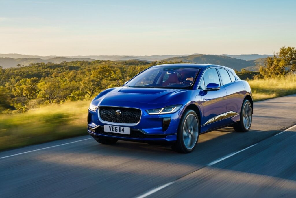 Blue electric car driving on scenic road with green hills and clear sky in the background.