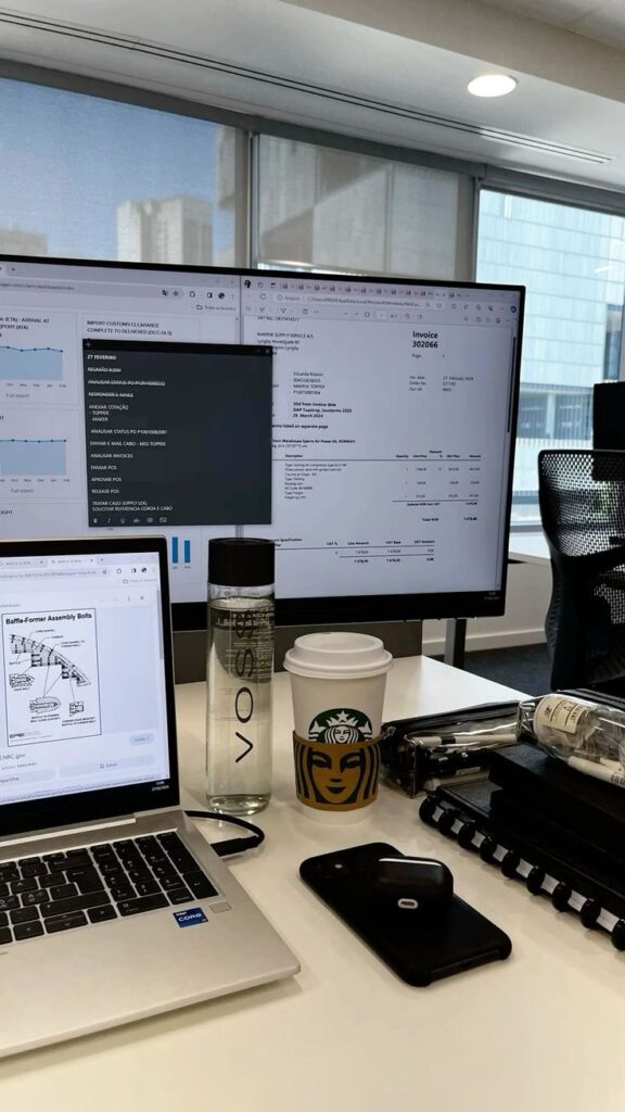 Office setup with a laptop, dual monitors displaying graphs and documents, coffee, water bottle, and phone on desk.