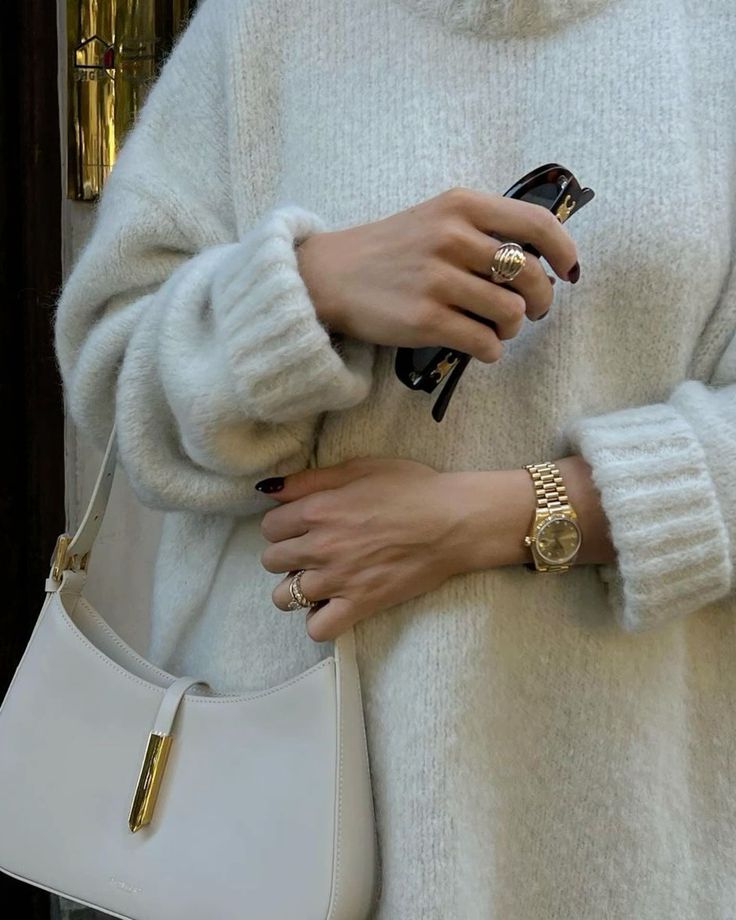 Stylish woman in cozy sweater holding handbag and sunglasses, wearing gold watch and rings.