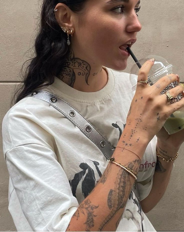 Woman with tattoos sipping iced drink through straw, wearing casual white shirt and accessories.