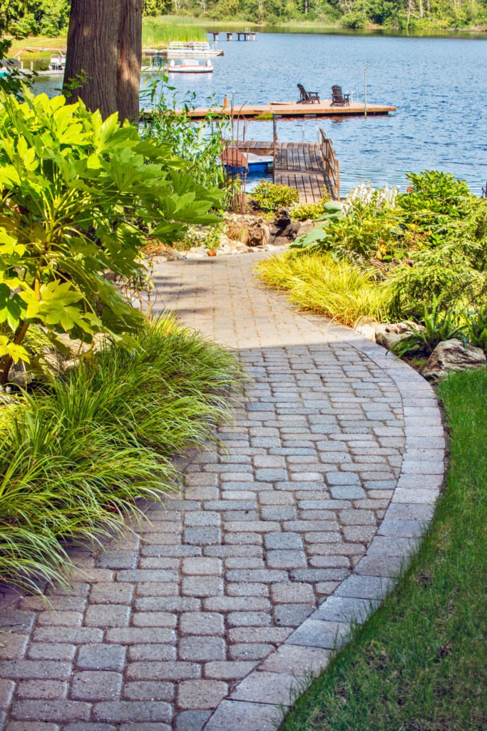 Scenic lakeside walkway with lush greenery leading to a wooden dock with chairs overlooking a tranquil blue lake.