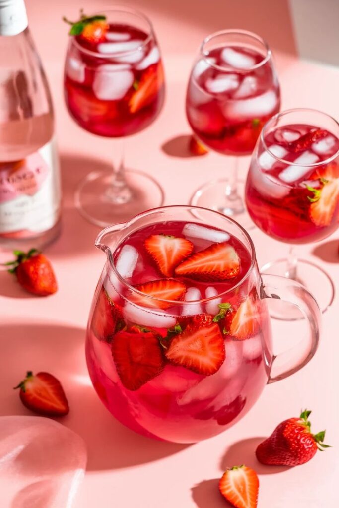 Refreshing strawberry sangria with ice in a pitcher and glasses on a pink background.