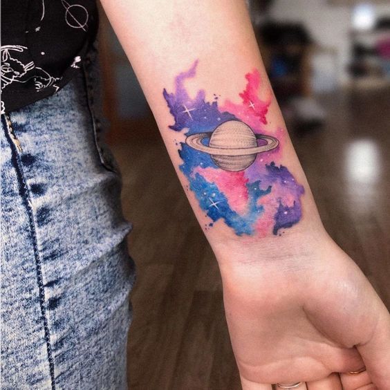 Colorful Saturn tattoo on forearm with galaxy background in pink, blue, and purple shades.